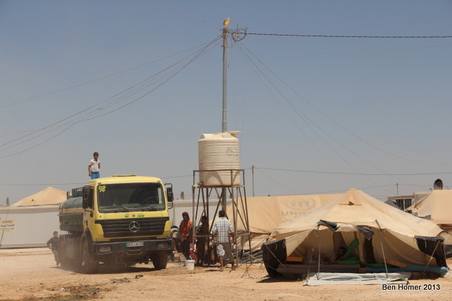 A truck delivers water to one of many elevated tanks in the camp. Behind it, an electricity pole with many additional lines coming off of it. Enterprising refugees charge five Jordanian Dollars to connect tents to the power grid.