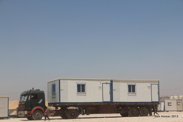 A truck delivers caravans to the cam. Caravans are highly sought over as compared to tents and there is a black market for these housing units. Flags on the outside of the caravan indicate the country which donated them. 